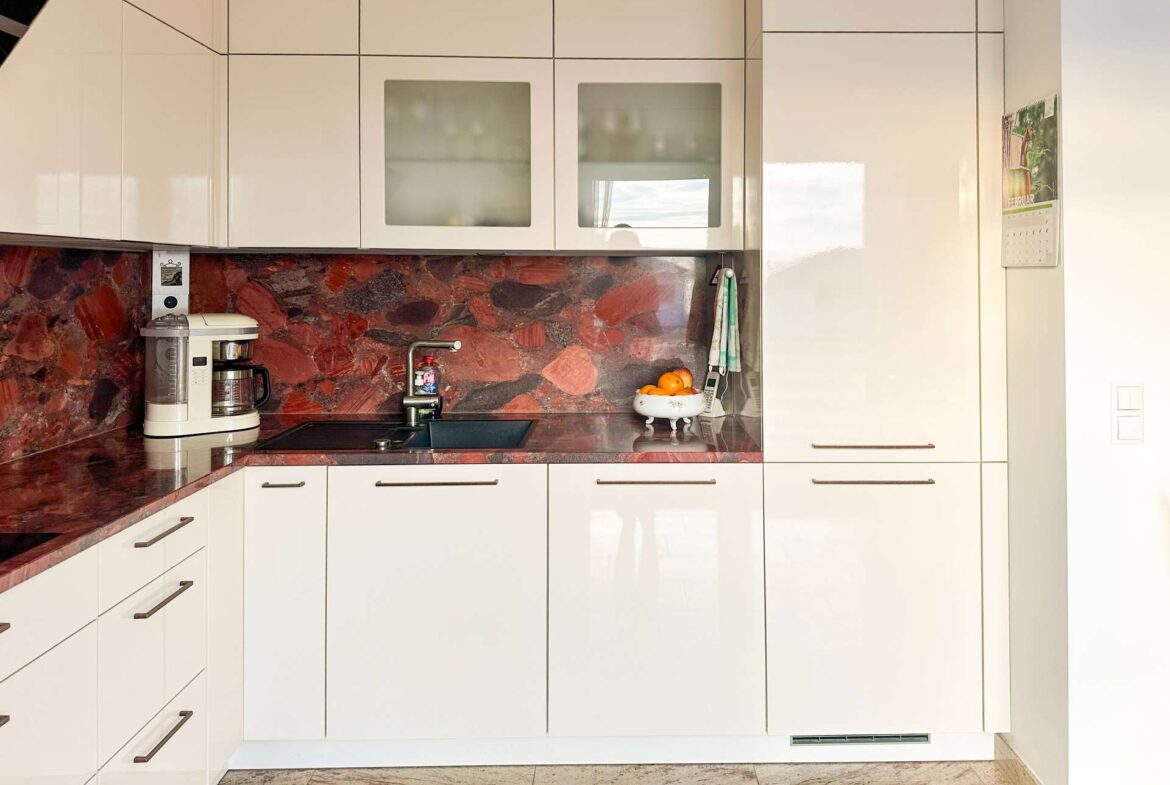 Modern white kitchen with red marble countertops and backsplash, upper and lower cabinets, and a sink area in the center left. A white coffee maker sits on the counter by the sink, with a fruit bowl and calendar on the right wall. The floor is tiled in light beige.