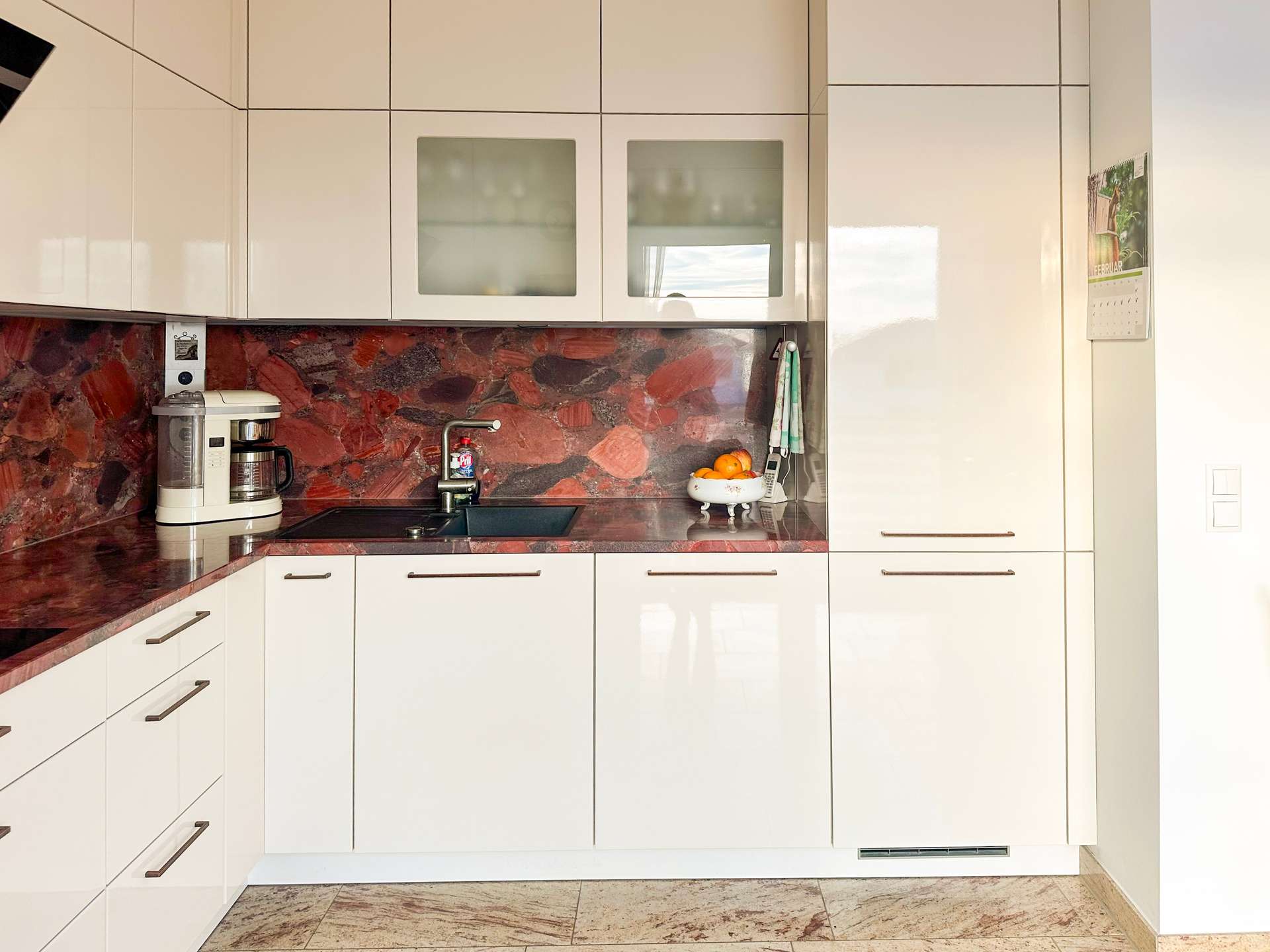 Modern white kitchen with red marble countertops and backsplash, upper and lower cabinets, and a sink area in the center left. A white coffee maker sits on the counter by the sink, with a fruit bowl and calendar on the right wall. The floor is tiled in light beige.