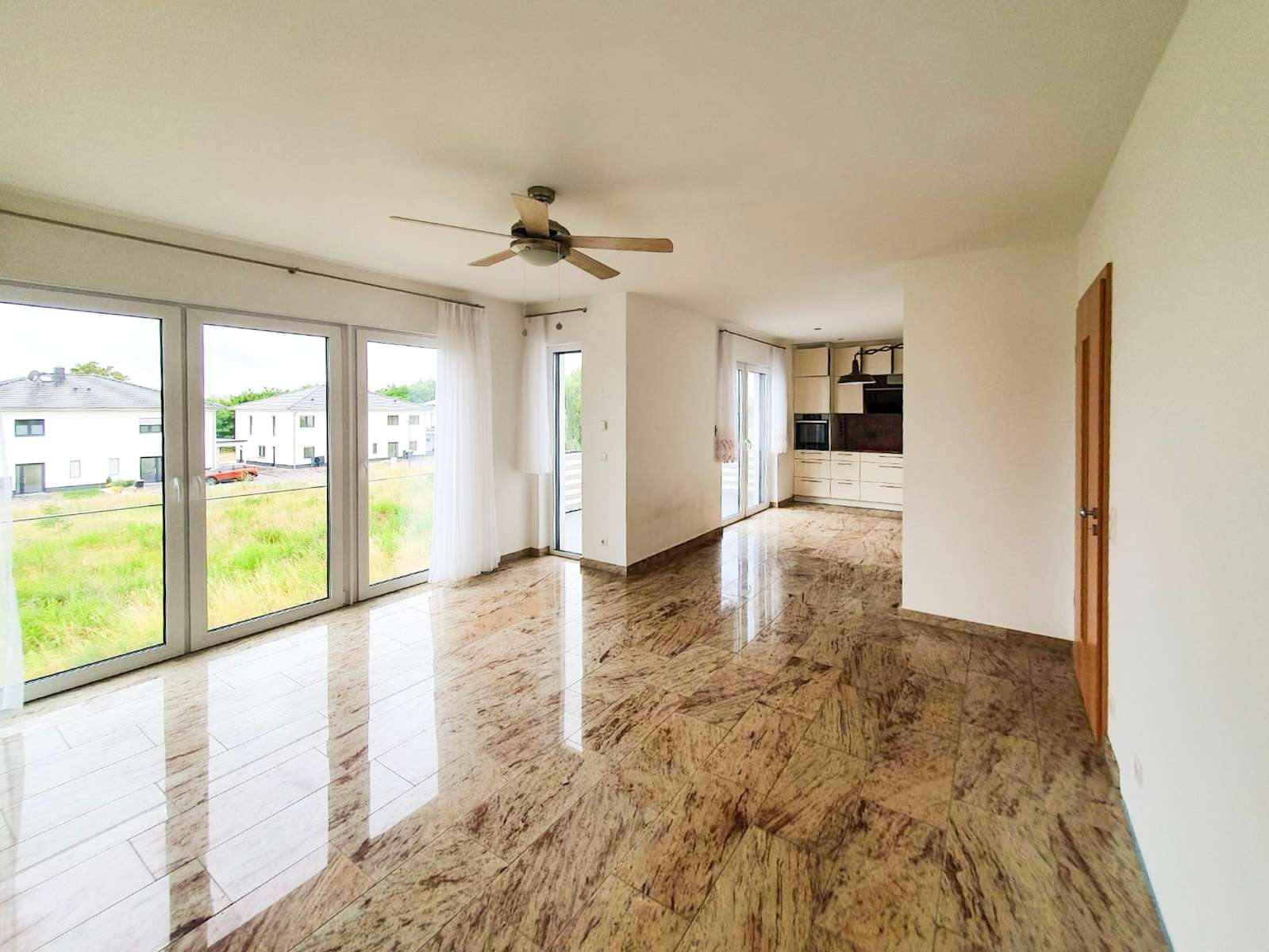 Open-plan living area with large glass doors to the outside, marble floors, and a ceiling fan, leading to a bright kitchen area in the back corner.