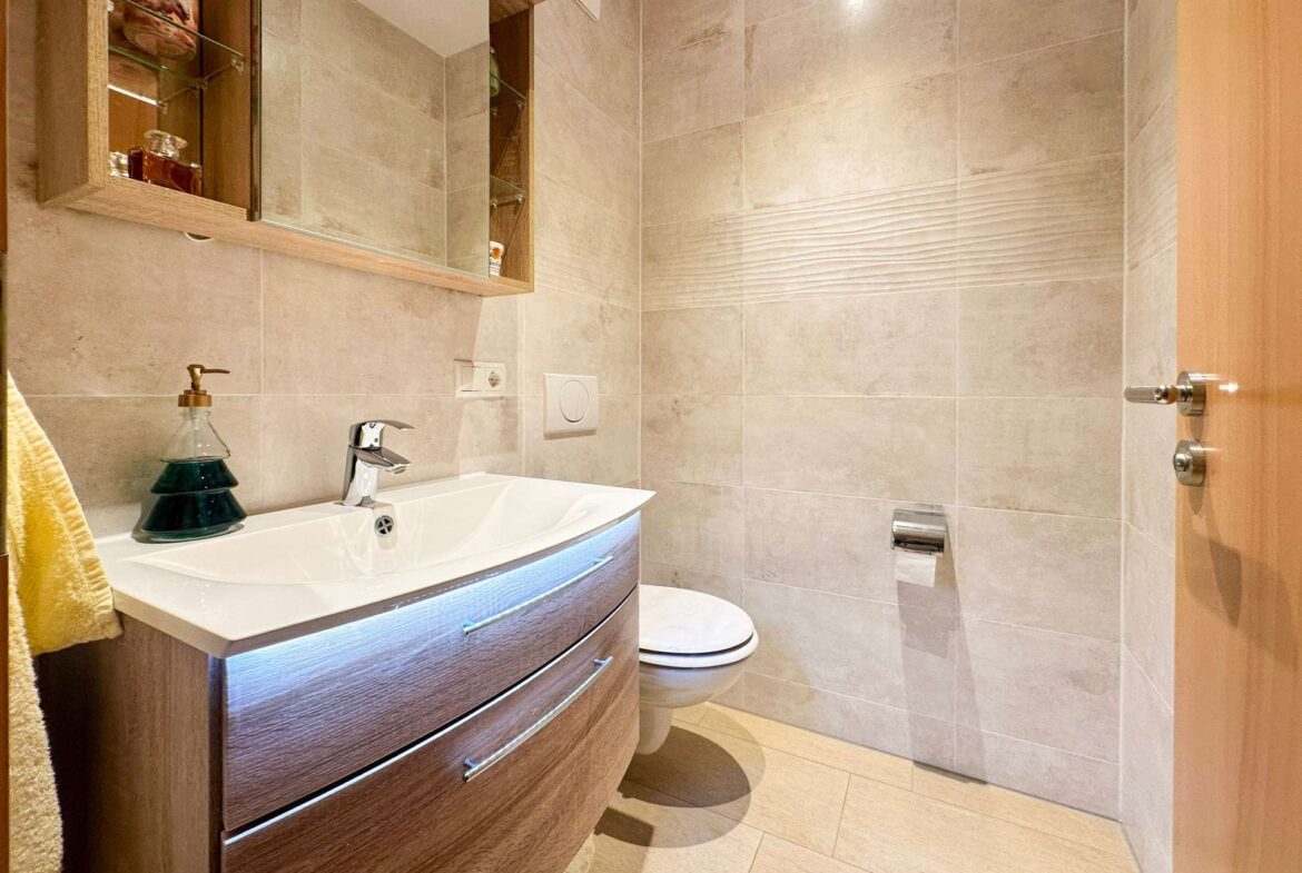 Modern bathroom with a curved wooden vanity, white sink, brass faucet, and mirrored cabinet against beige tiled walls with a yellow towel on the left.