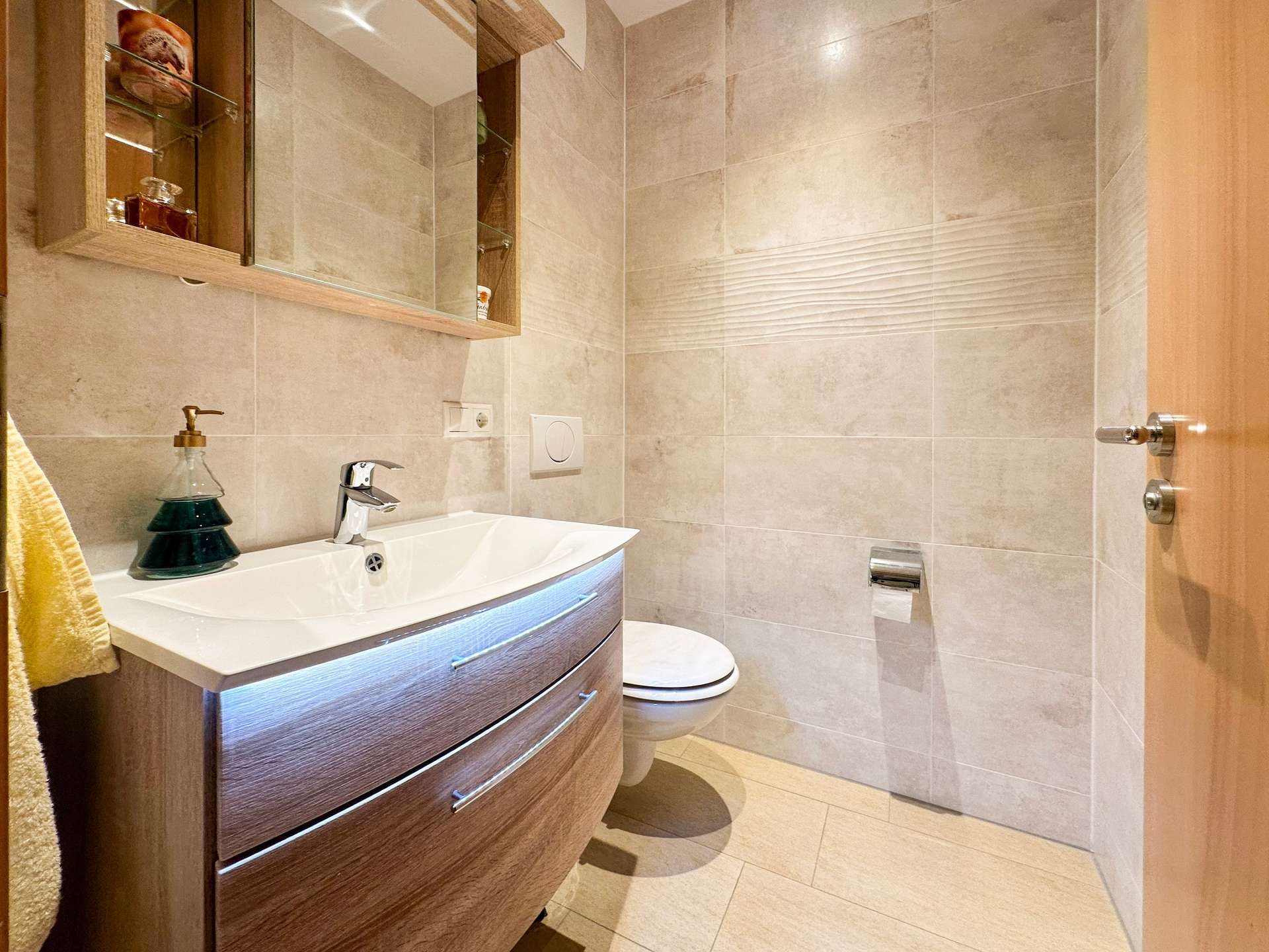 Modern bathroom featuring a curved wooden vanity with white basin, wall mirror cabinet, and beige tiled walls and floor.