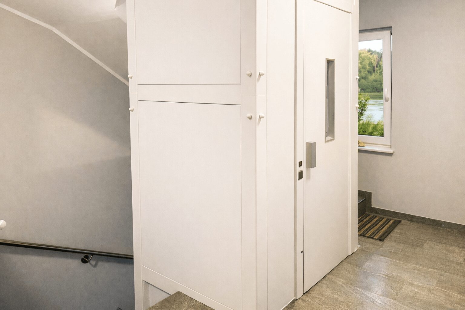 White built-in storage cabinets line a hallway, with stairs on the left and a window at the end showing greenery outside.