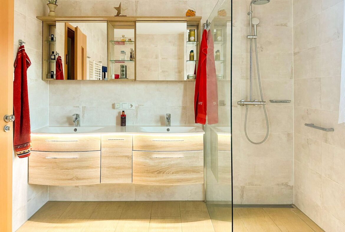Modern bathroom with a light wood double-sink vanity, large mirrored medicine cabinet, and glass-enclosed shower with chrome fixtures.
