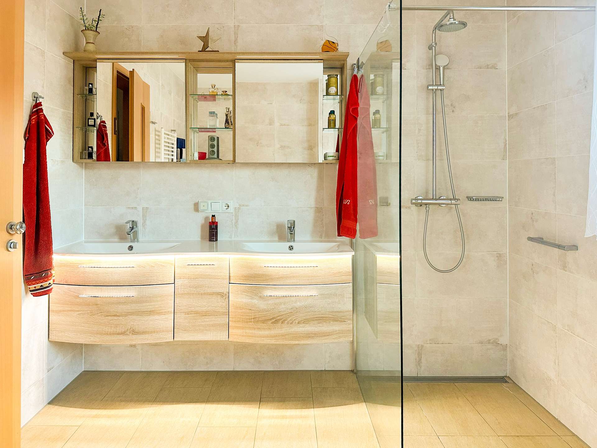 Modern bathroom with a light wood double-sink vanity, large mirrored medicine cabinet, and glass-enclosed shower with chrome fixtures.