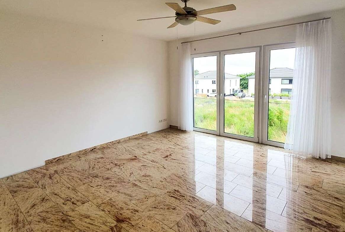 Empty living room with marble-like tiled floor, white walls, a ceiling fan, and large glass doors opening to a yard with houses outside.