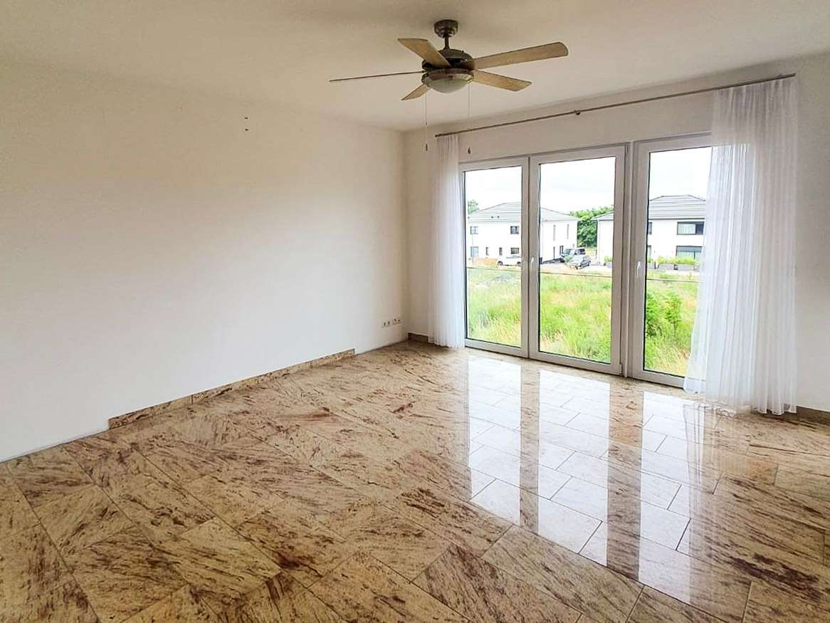 Empty living room with marble-like tiled floor, white walls, a ceiling fan, and large glass doors opening to a yard with houses outside.