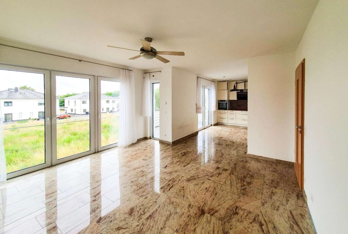 Bright open-plan living area with large glass doors to the outdoors, marble floors, and a ceiling fan; kitchen visible in the back.
