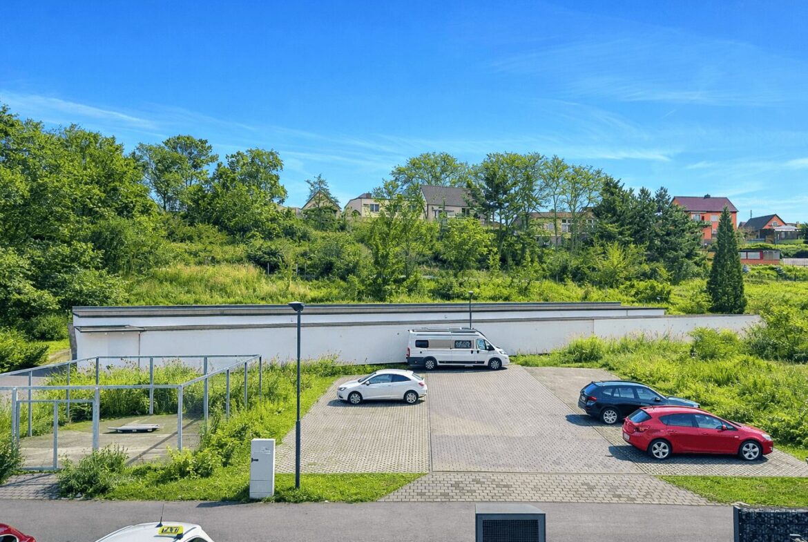 Parking lot with a white van and several cars beside a grassy embankment and a low white building, trees and houses beyond under a blue sky.