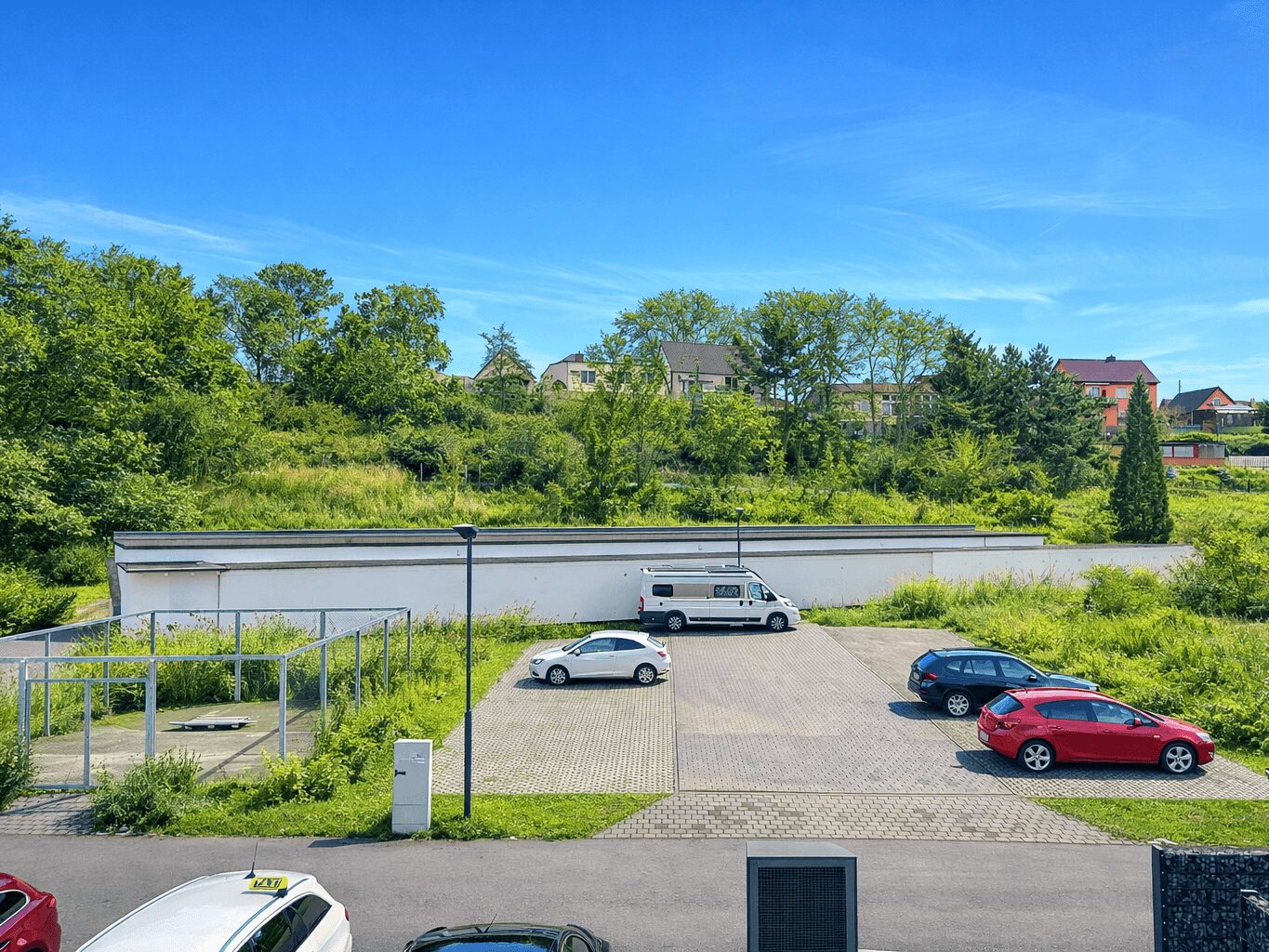 Parking lot with a white van and several cars beside a grassy embankment and a low white building, trees and houses beyond under a blue sky.