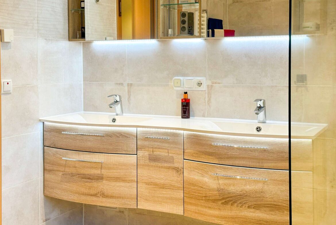 Double-sink bathroom vanity with light wood drawers, a white countertop, and chrome faucets beneath a large mirror with under-cabinet LED lighting; glass shower divider on the right side.