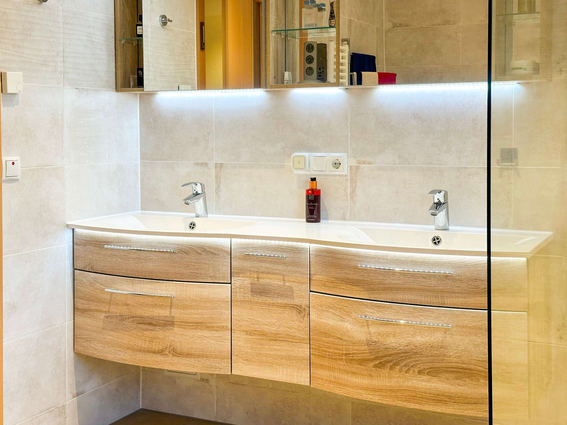 Double-sink bathroom vanity with light wood drawers, a white countertop, and chrome faucets beneath a large mirror with under-cabinet LED lighting; glass shower divider on the right side.