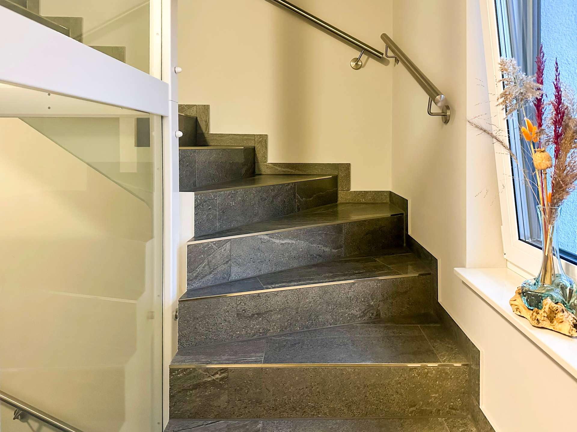 Indoor modern staircase with dark stone steps, glass railing on the left, and a window with dried-flower arrangement on the right.