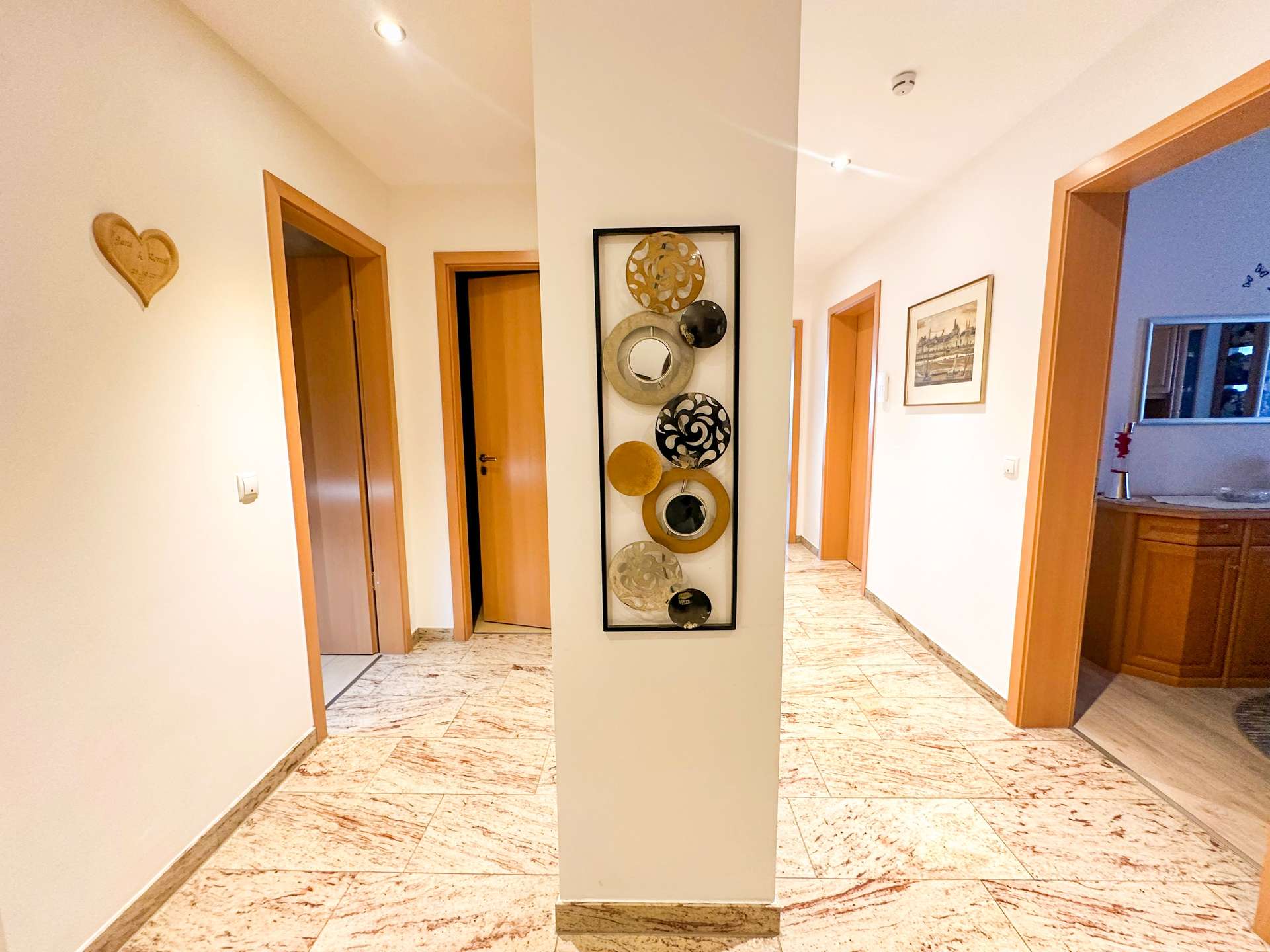 Bright hallway with marble floors, wooden doors, and a tall central pillar displaying circular metal art pieces.