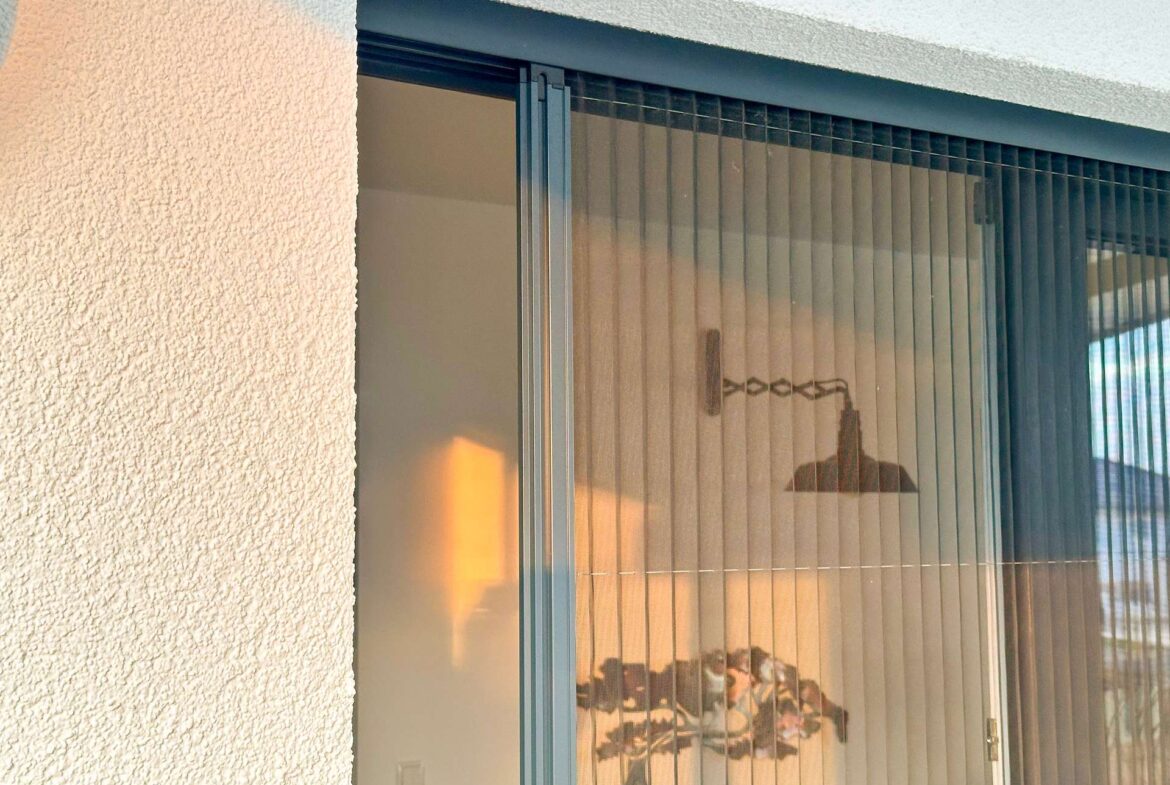 Exterior sliding glass door with vertical blinds, glimpses of a lit living room inside, and a textured stucco wall nearby.