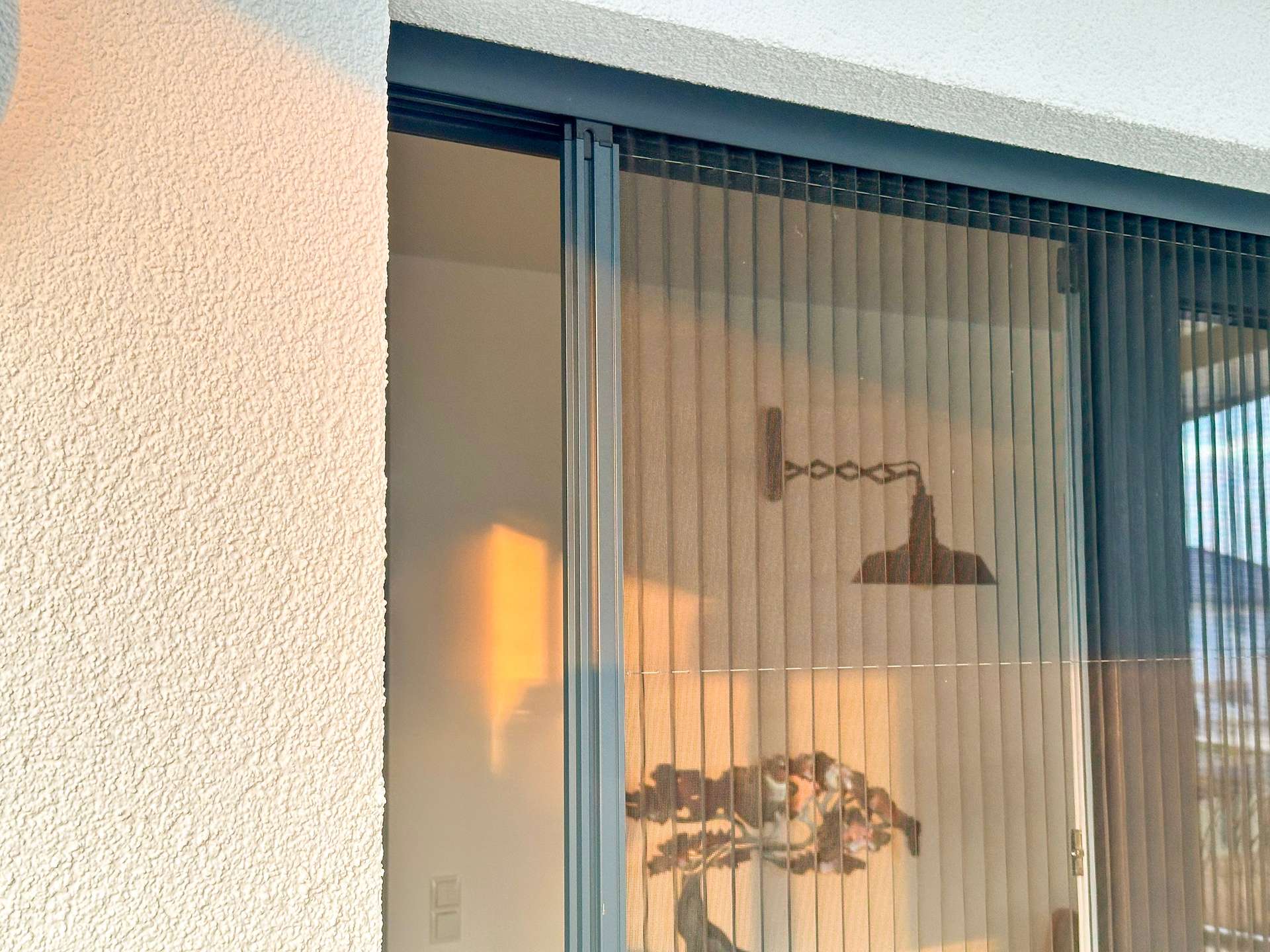 Exterior sliding glass door with vertical blinds, glimpses of a lit living room inside, and a textured stucco wall nearby.