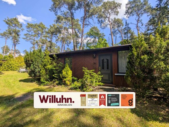 Brown wooden house with a dark green door, surrounded by tall trees and a Willuhn Immobilien sign in the foreground.