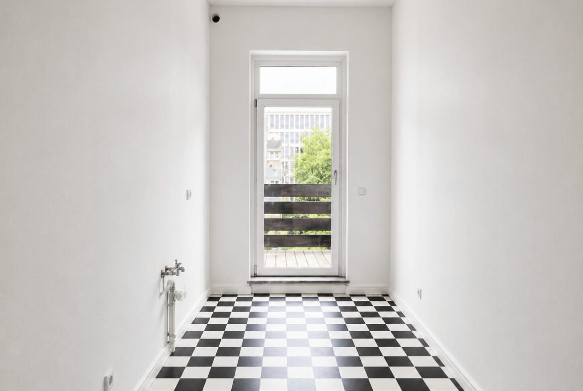 Narrow white room with a glass door opening to a balcony, featuring a black-and-white checkerboard floor and a view of trees outside.