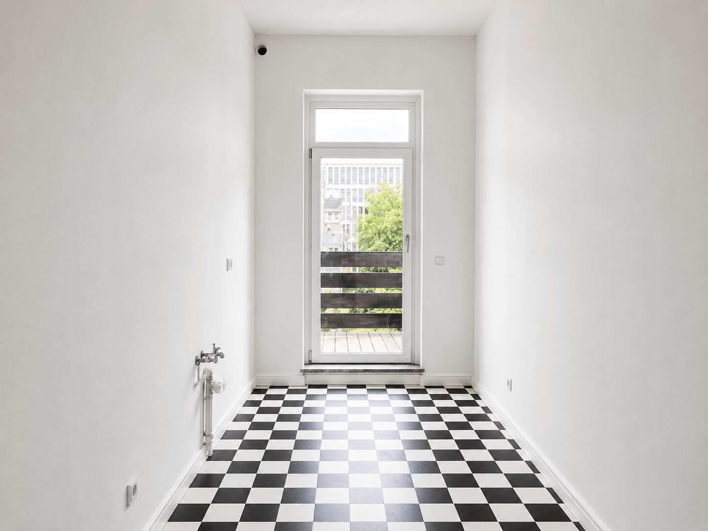 Narrow white room with a glass door opening to a balcony, featuring a black-and-white checkerboard floor and a view of trees outside.