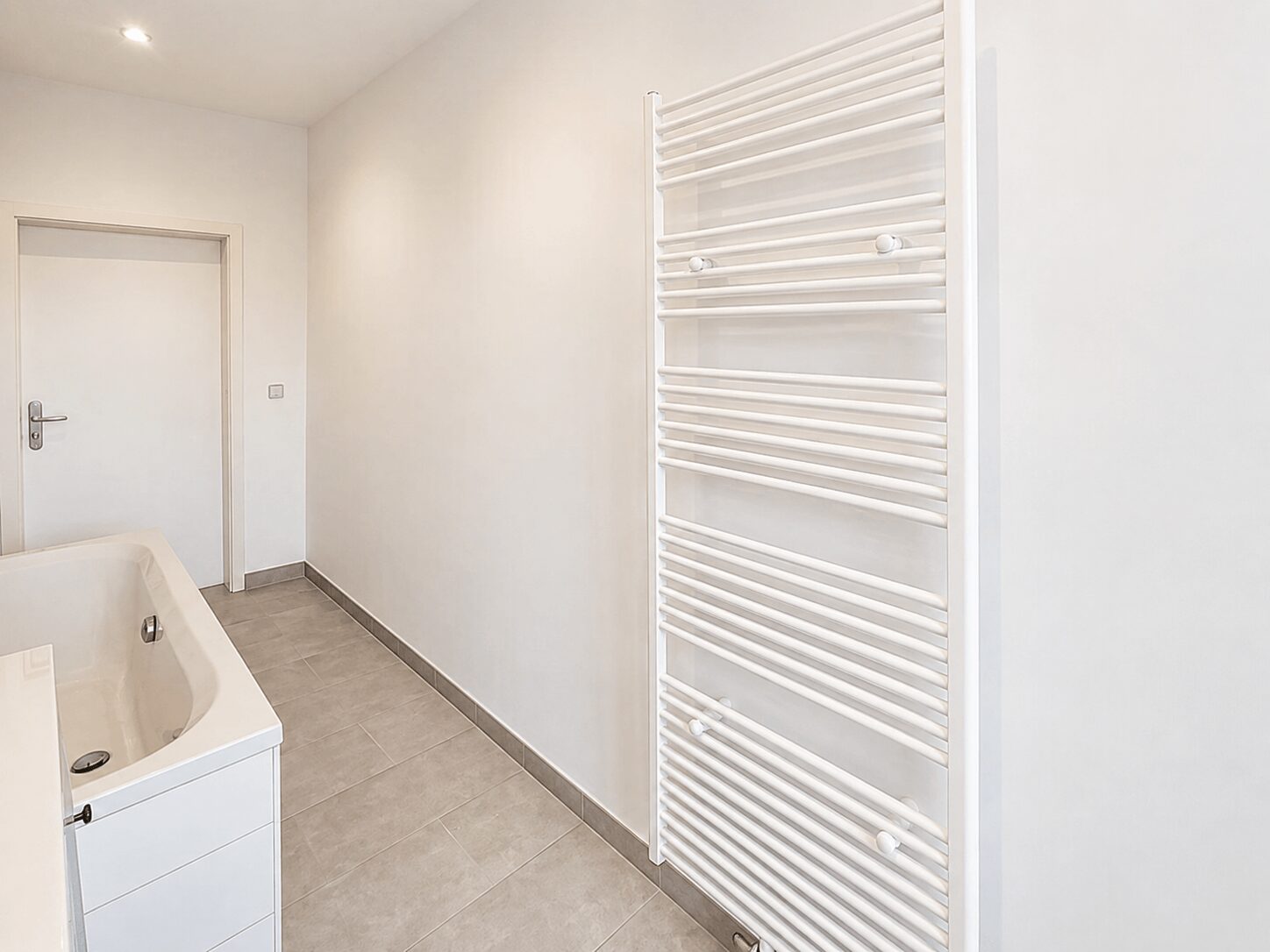 Bright white bathroom with a wall-mounted towel warmer, a white vanity with sink, gray floor tiles, and a closed door.