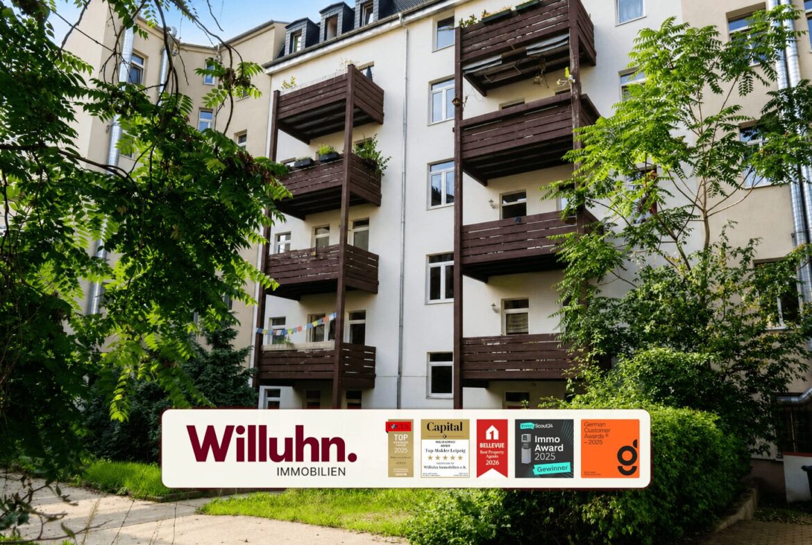 Apartment building with dark wooden balconies, surrounded by green trees, and a large Willuhn Immobilien sign in the foreground.