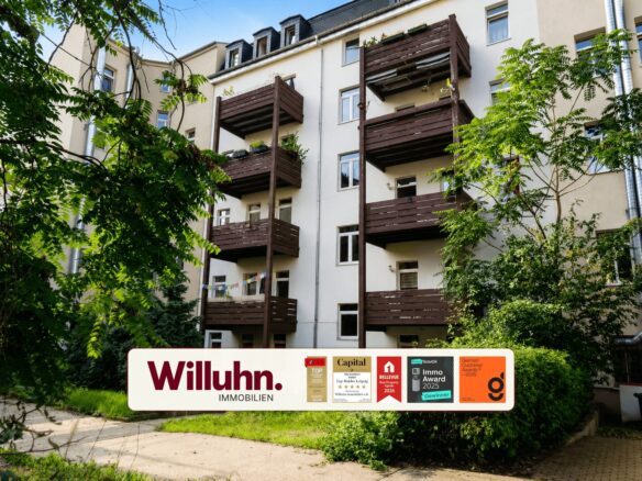 Apartment building with dark wooden balconies, surrounded by green trees, and a large Willuhn Immobilien sign in the foreground.