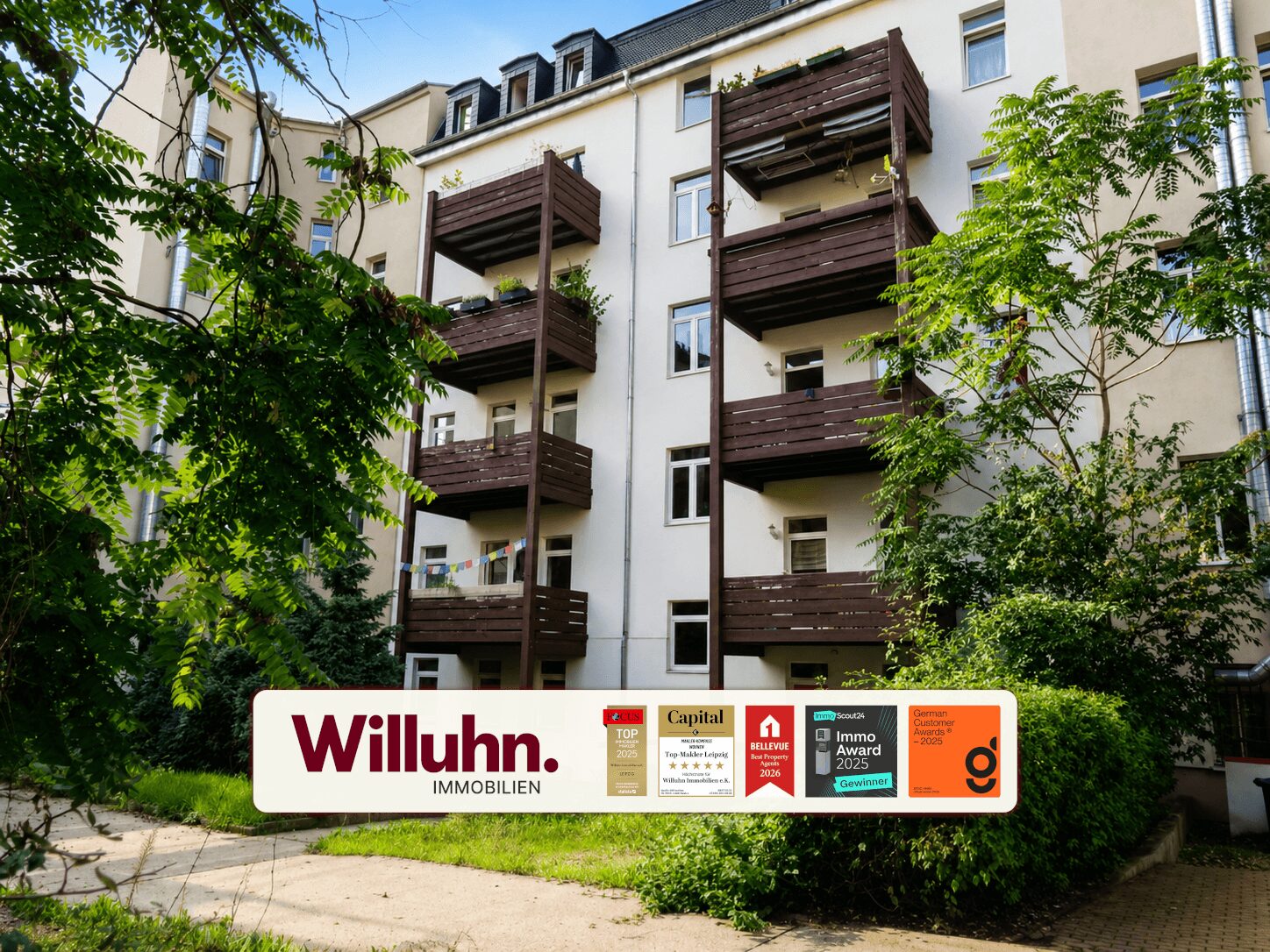 Apartment building with dark wooden balconies, surrounded by green trees, and a large Willuhn Immobilien sign in the foreground.