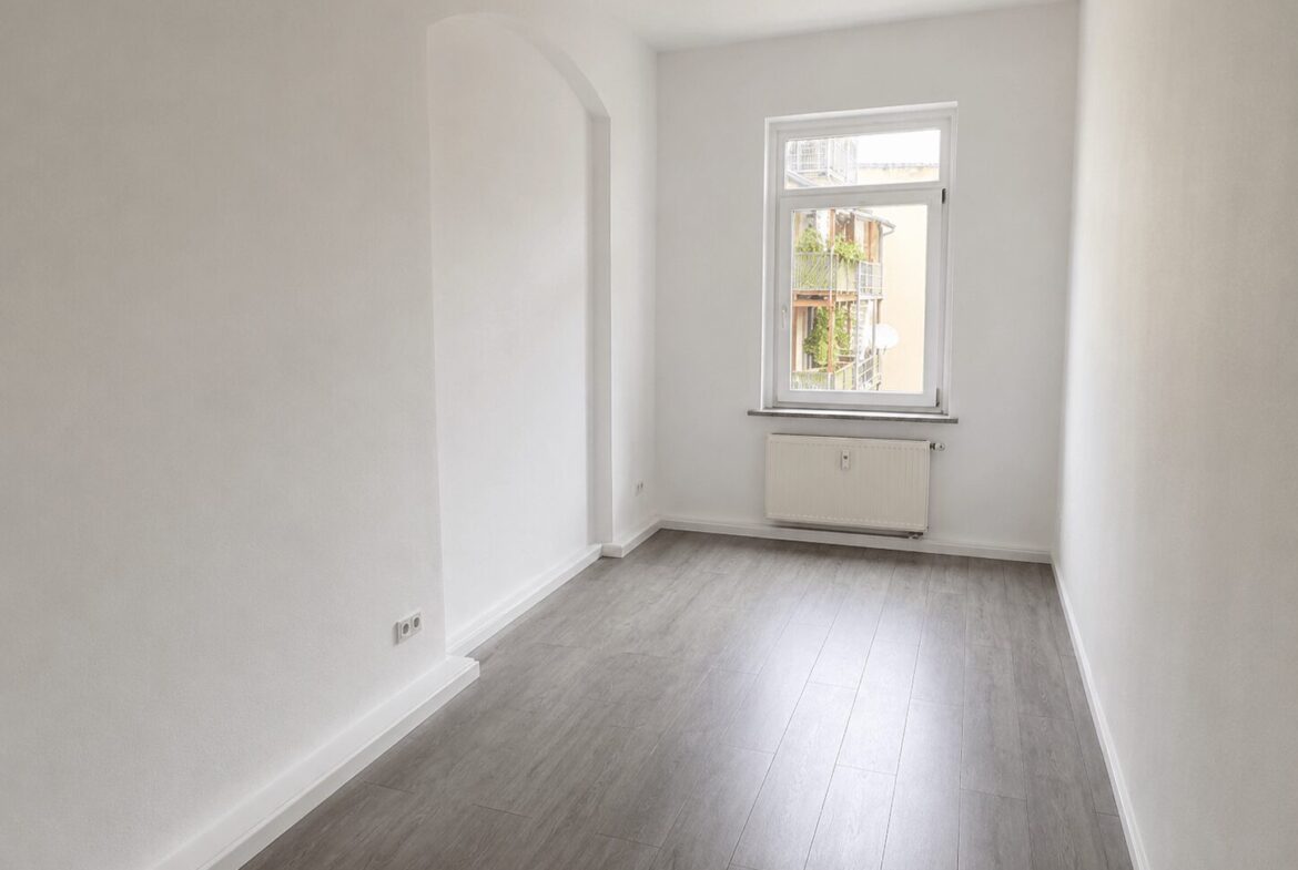 Bright, empty white room with a single window, radiator under the window, and light gray wood-look flooring.