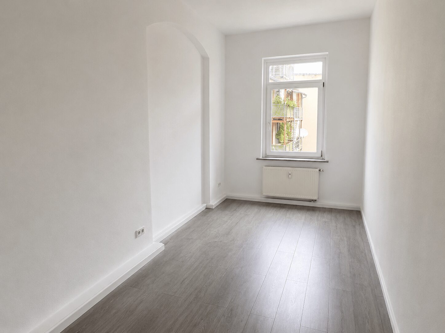 Bright, empty white room with a single window, radiator under the window, and light gray wood-look flooring.