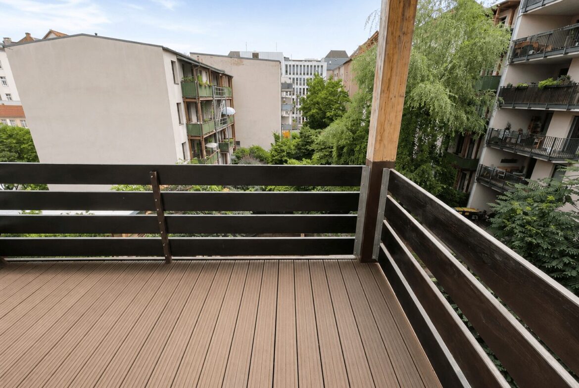View from a wooden balcony on a sunny day, looking out over neighboring apartment buildings and green trees in a courtyard. The deck has brown planks and dark railing with a vertical wooden post nearby.