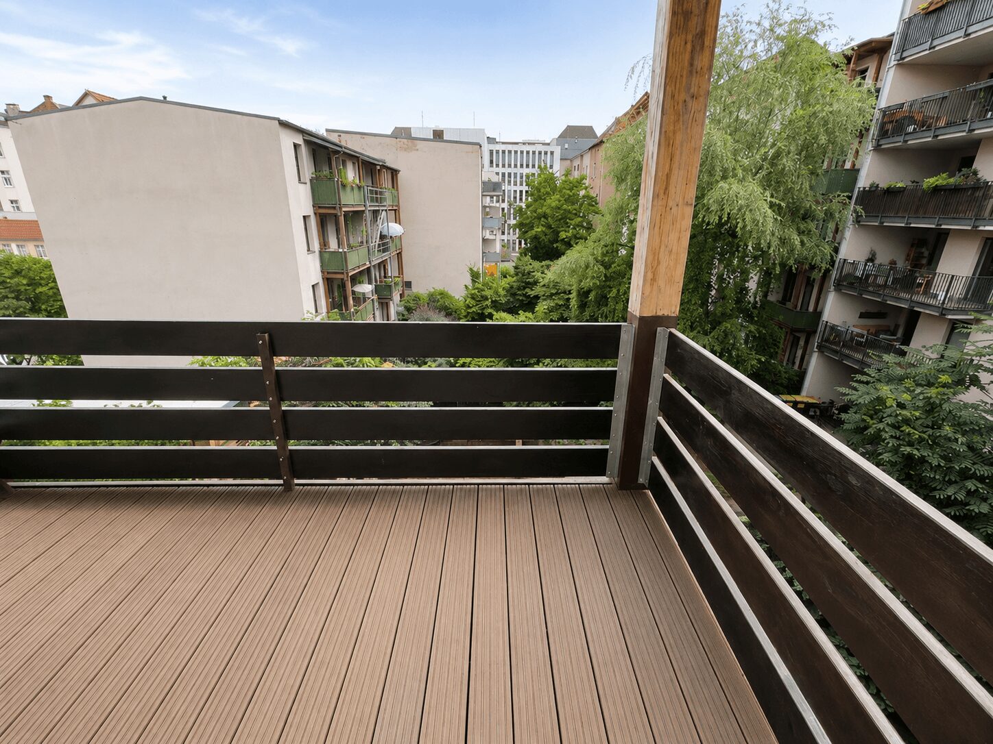View from a wooden balcony on a sunny day, looking out over neighboring apartment buildings and green trees in a courtyard. The deck has brown planks and dark railing with a vertical wooden post nearby.