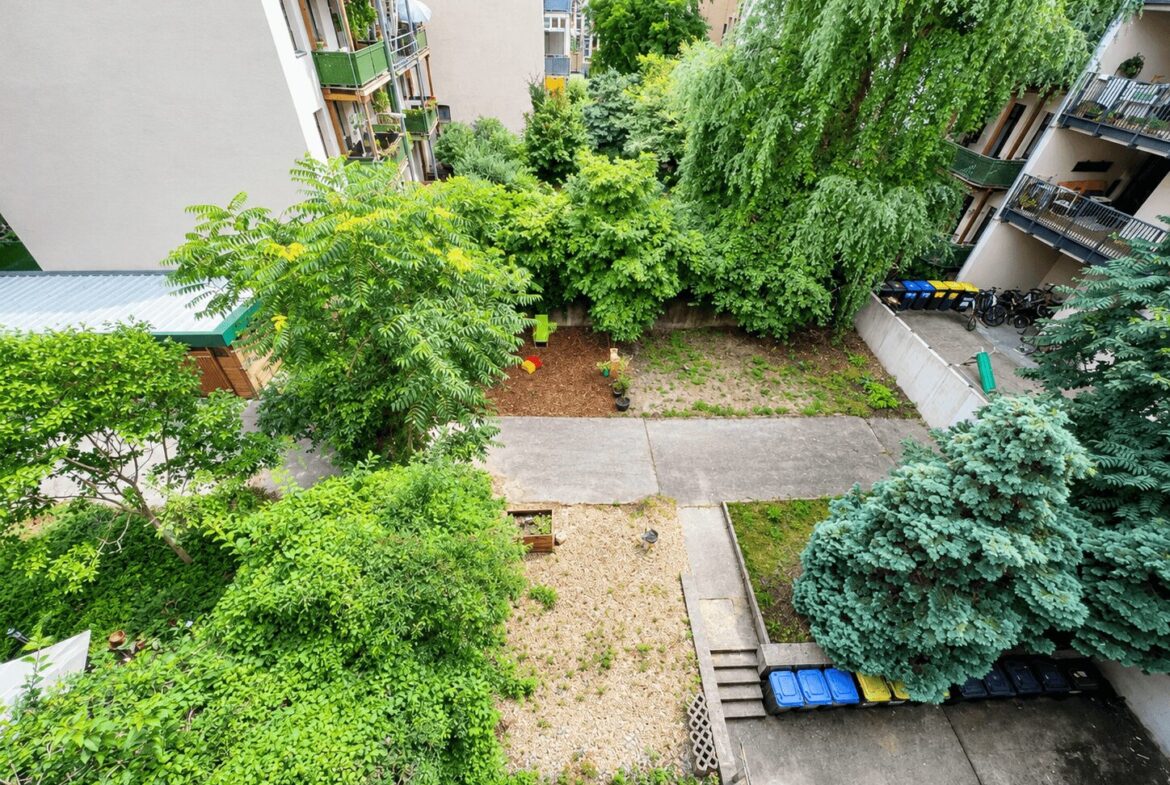 Bird's-eye view of a courtyard between apartment buildings with dense trees, shrubs, and a few bikes and recycling bins along the edge.