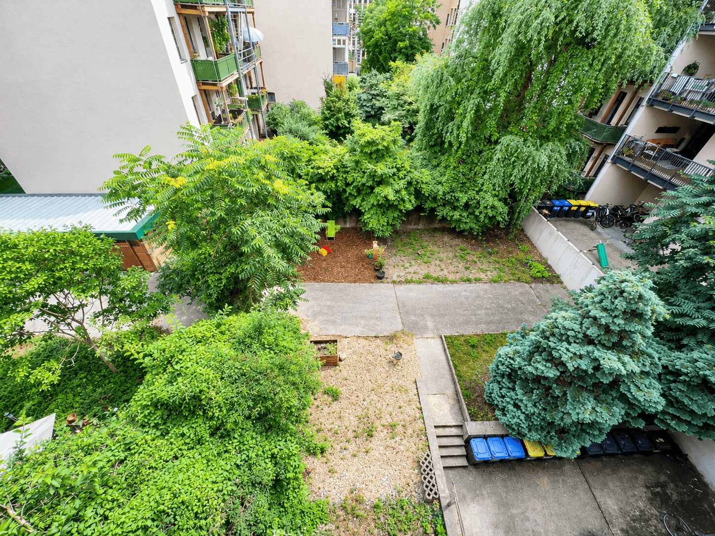 Bird's-eye view of a courtyard between apartment buildings with dense trees, shrubs, and a few bikes and recycling bins along the edge.