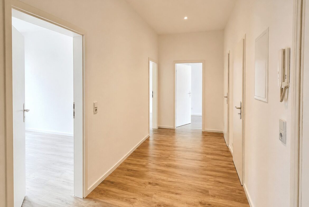 Bright, minimalist hallway with light wood flooring and white walls; several closed white doors along both sides.