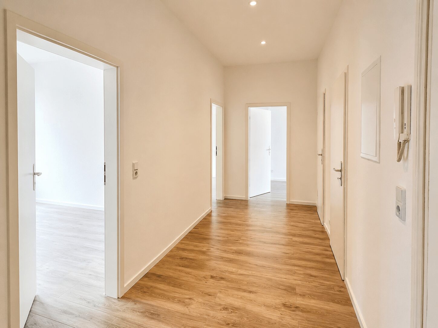 Bright, minimalist hallway with light wood flooring and white walls; several closed white doors along both sides.