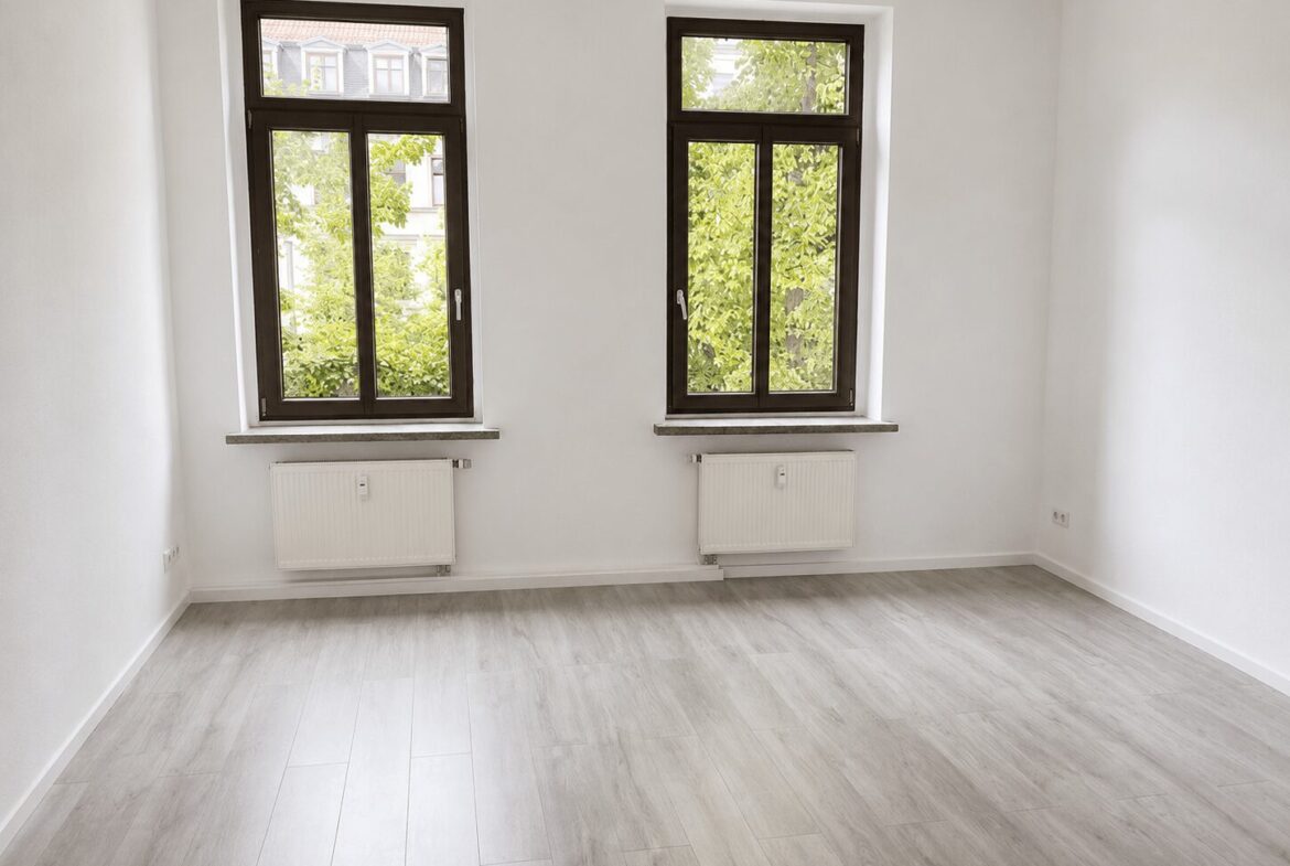Bright empty room with two tall dark-framed windows, white walls, light gray wood flooring, and radiators beneath the windows; greenery visible outside. (Interior, unfurnished)