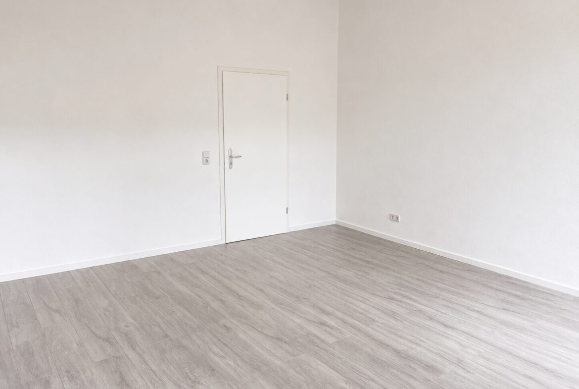 Empty white-walled room with light gray wood flooring, a closed white door on the left, and two electrical outlets on the right wall.