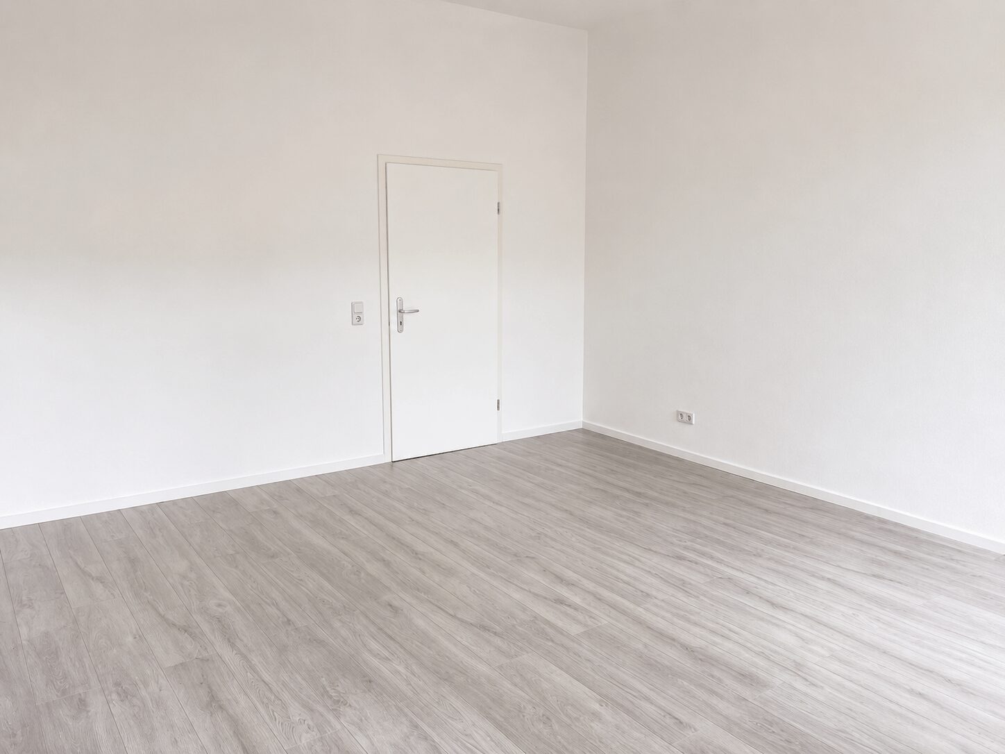 Empty white-walled room with light gray wood flooring, a closed white door on the left, and two electrical outlets on the right wall.