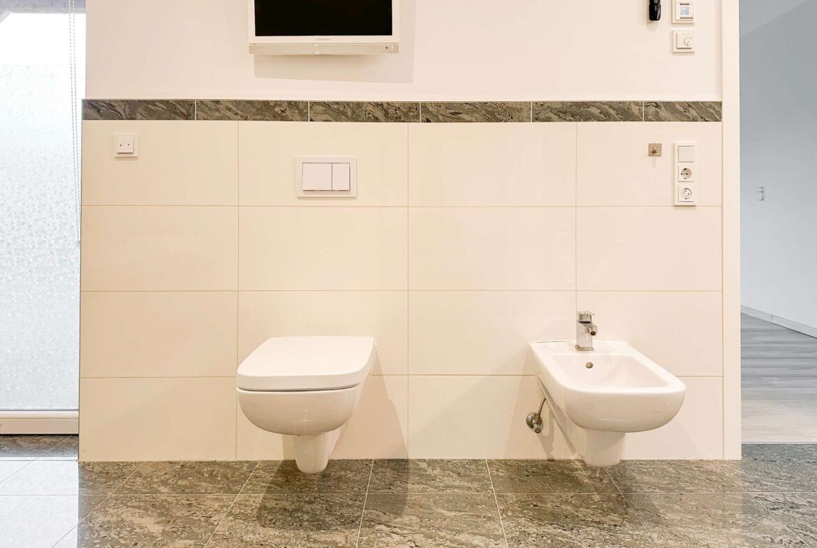 Modern bathroom with wall-mounted toilet and bidet, beige tiled walls, marble floor, and a small wall TV above.