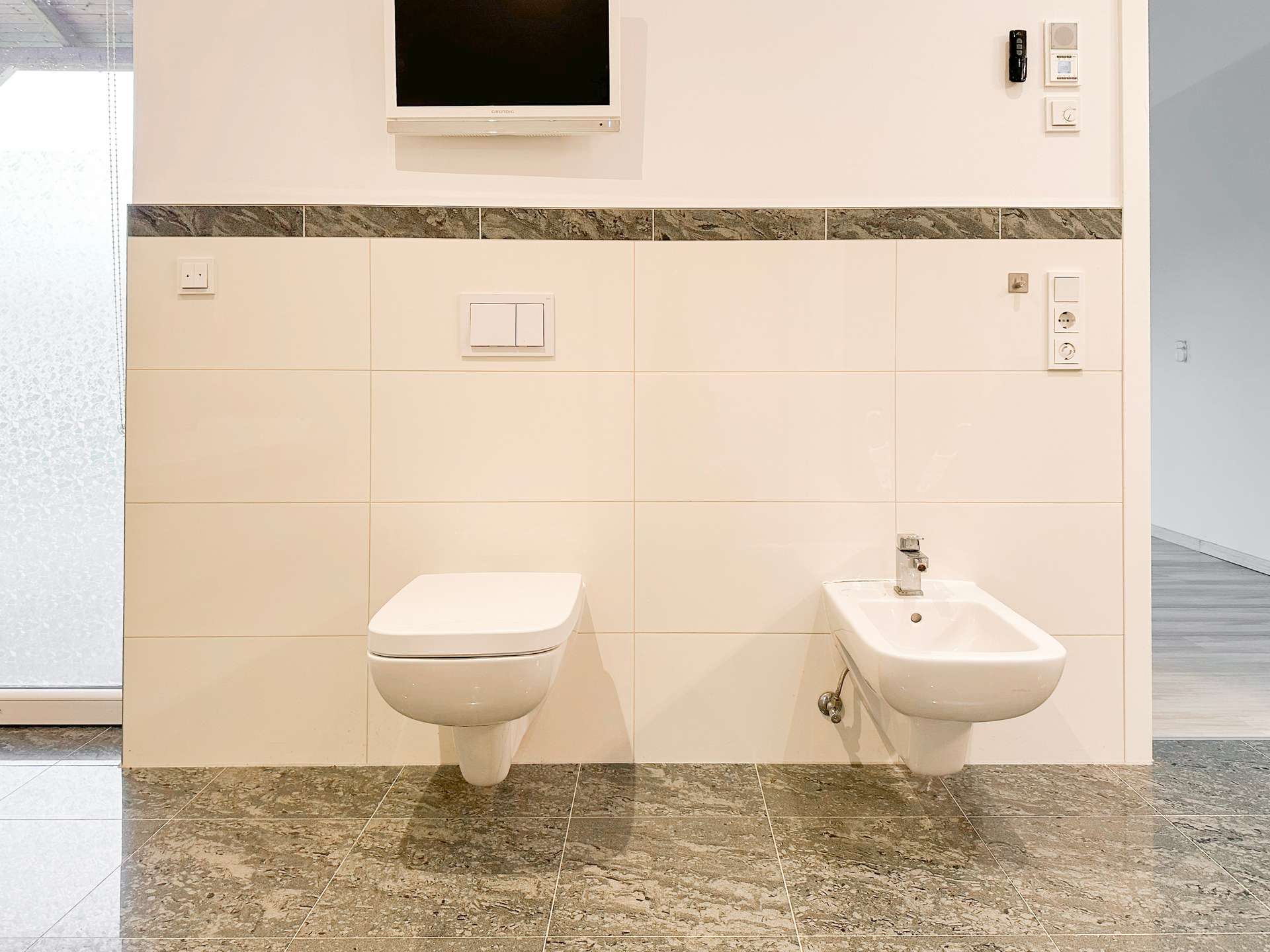 Modern bathroom with wall-mounted toilet and bidet, beige tiled walls, marble floor, and a small wall TV above.