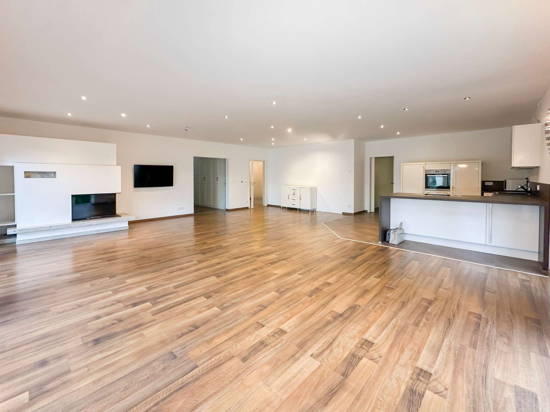 Spacious open-plan living area with hardwood floors, white walls, and a modern kitchen on the right.