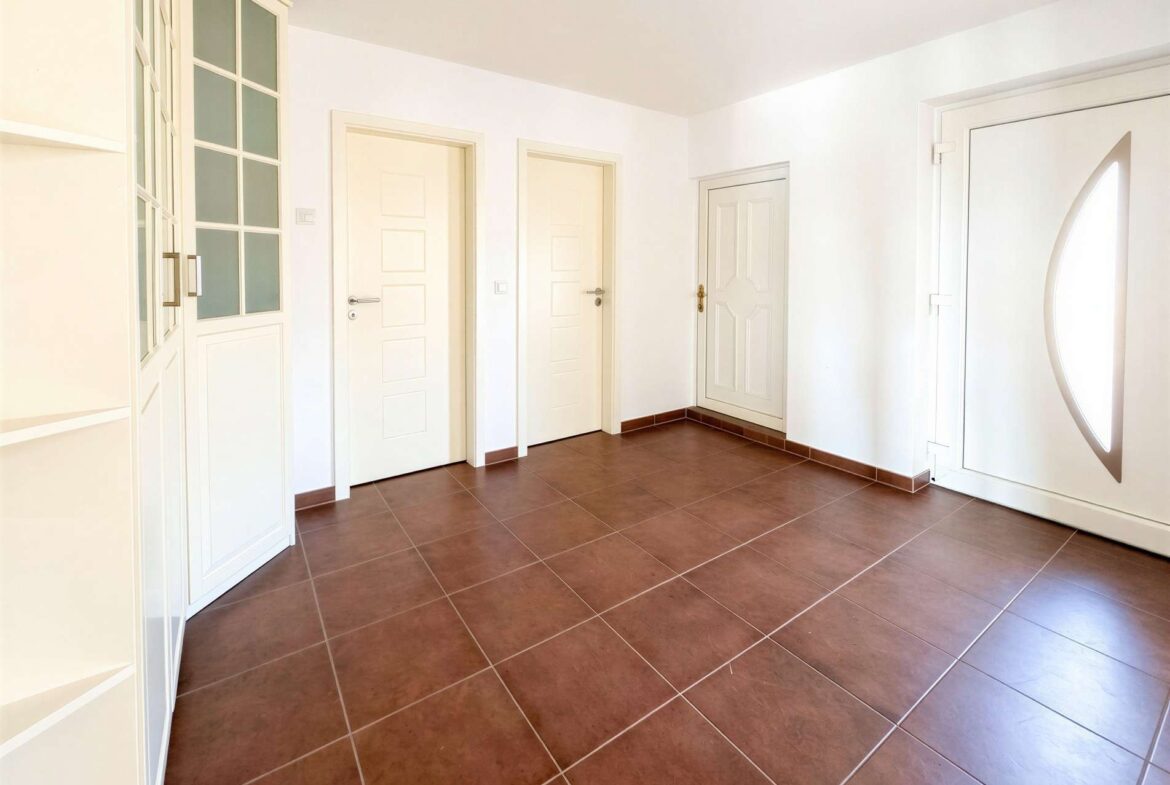 Bright hallway with white doors along a white wall, a tall glass-front cabinet on the left, and a door with an oval window on the right; brown tiled floor.