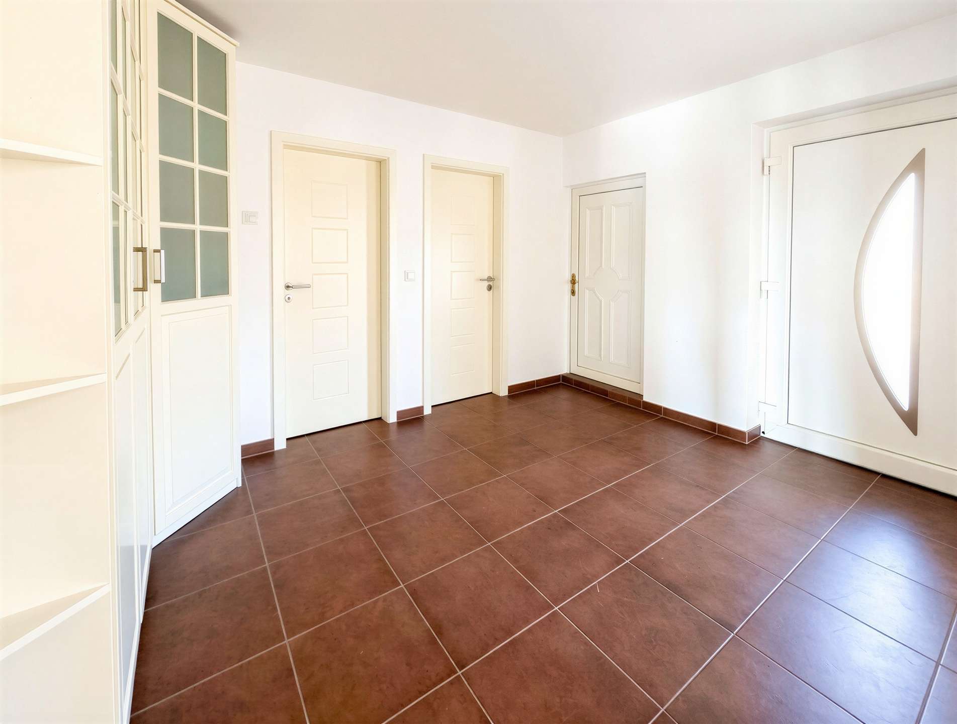 Bright hallway with white doors along a white wall, a tall glass-front cabinet on the left, and a door with an oval window on the right; brown tiled floor.