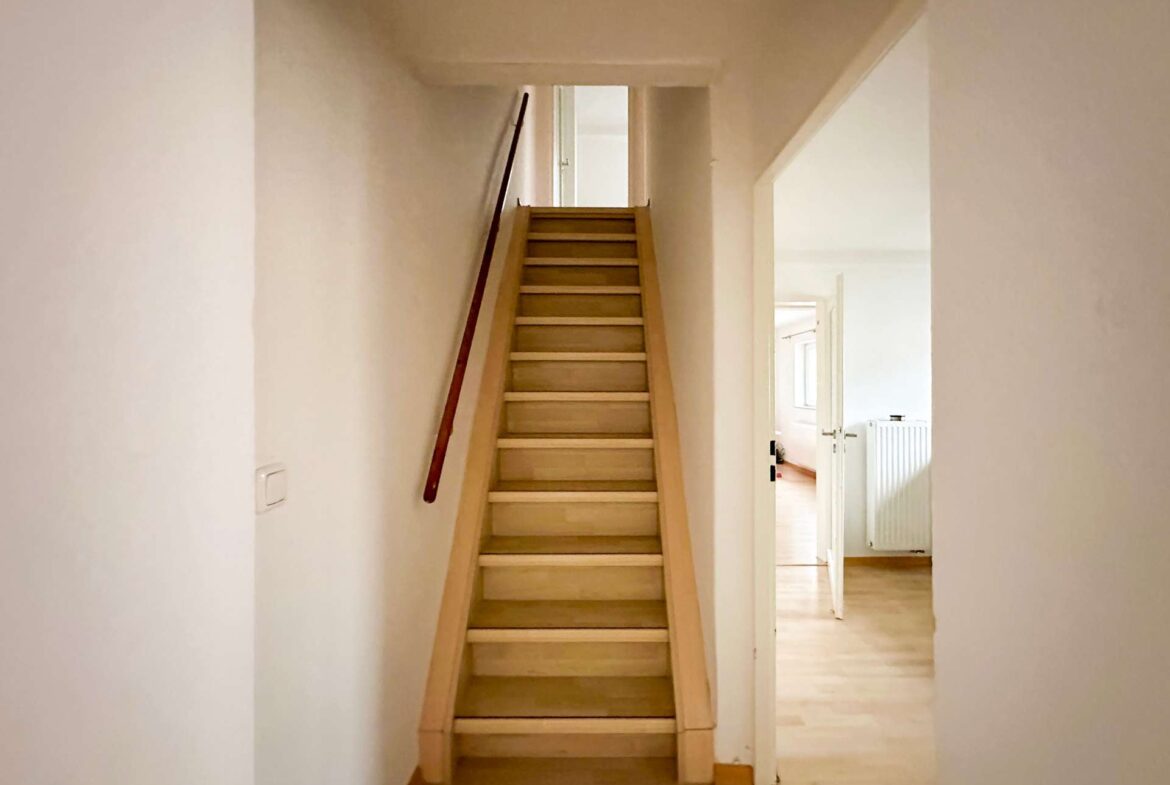 Wooden staircase in a narrow, cream-colored hallway leading up to a landing with an open doorway at the top.