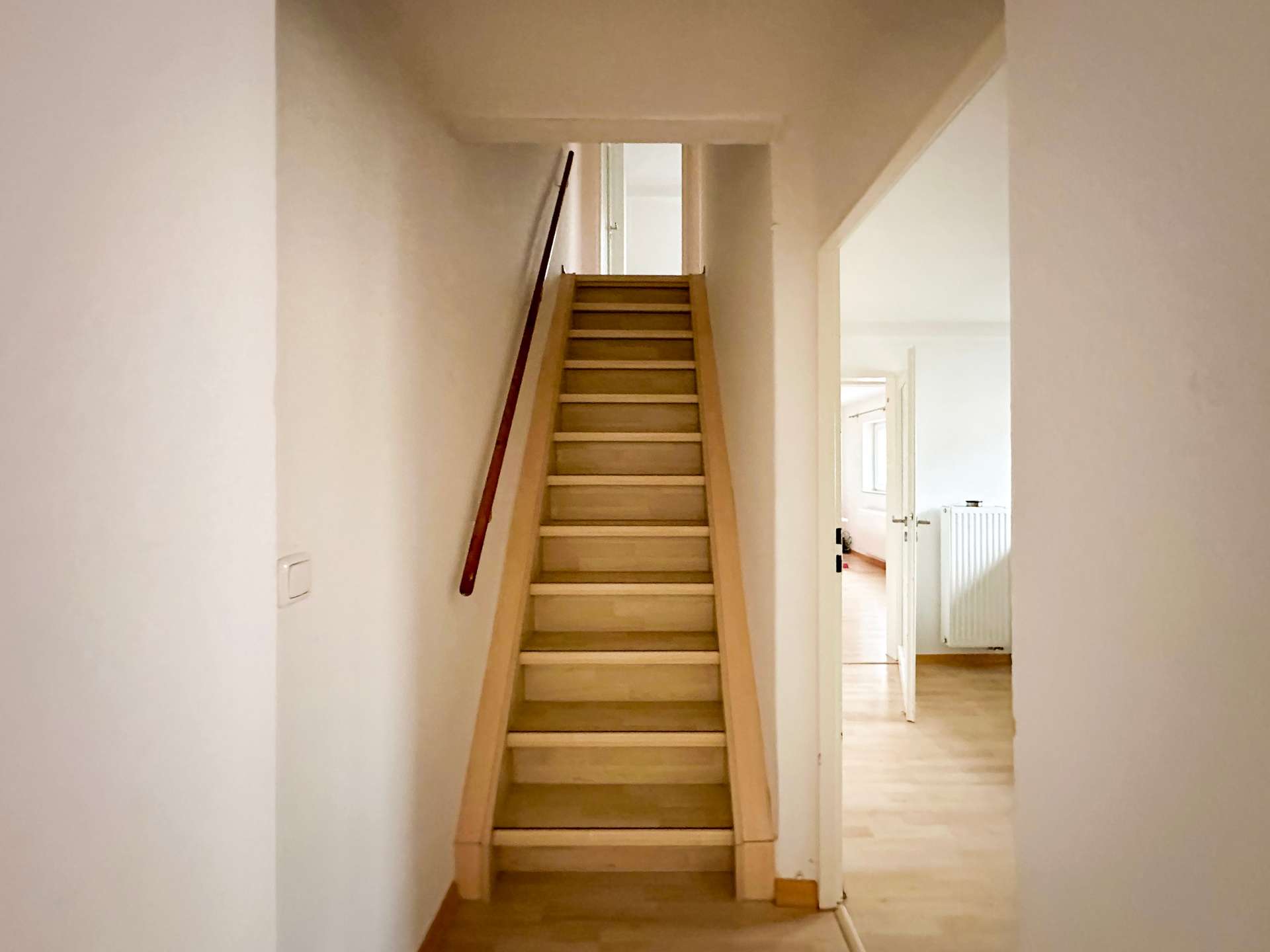 Wooden staircase in a narrow, cream-colored hallway leading up to a landing with an open doorway at the top.