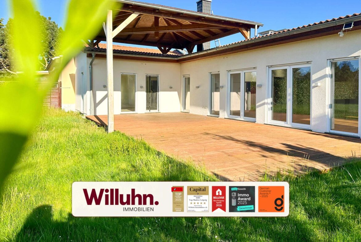 Modern single-story house with a wooden patio and large glass doors; foreground shows a Willuhn Immobilien real estate sign.