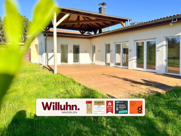Modern single-story house with a wooden patio and large glass doors; foreground shows a Willuhn Immobilien real estate sign.