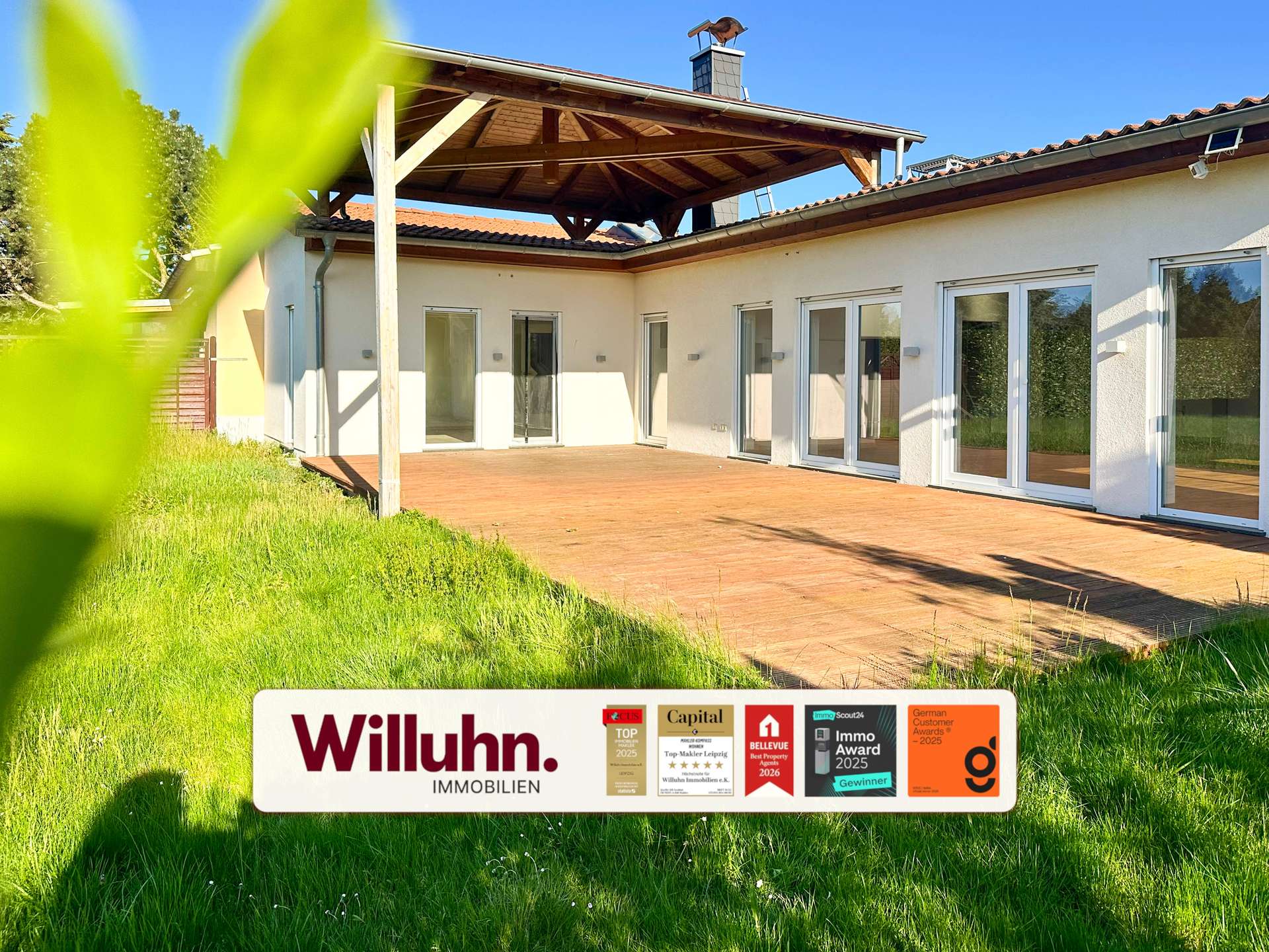 Modern single-story house with a wooden patio and large glass doors; foreground shows a Willuhn Immobilien real estate sign.