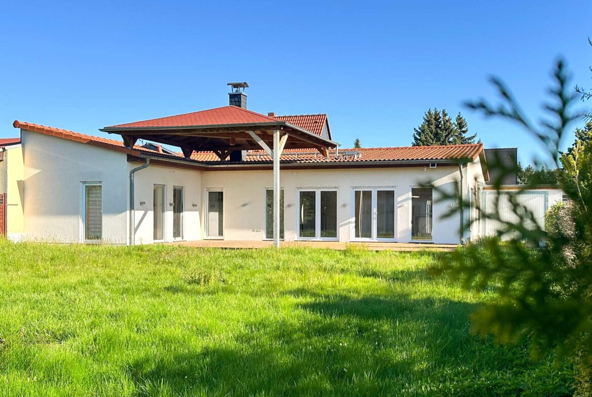 Single-story white house with a red-tiled roof and large glass doors, set in a sunny green yard under a clear blue sky.