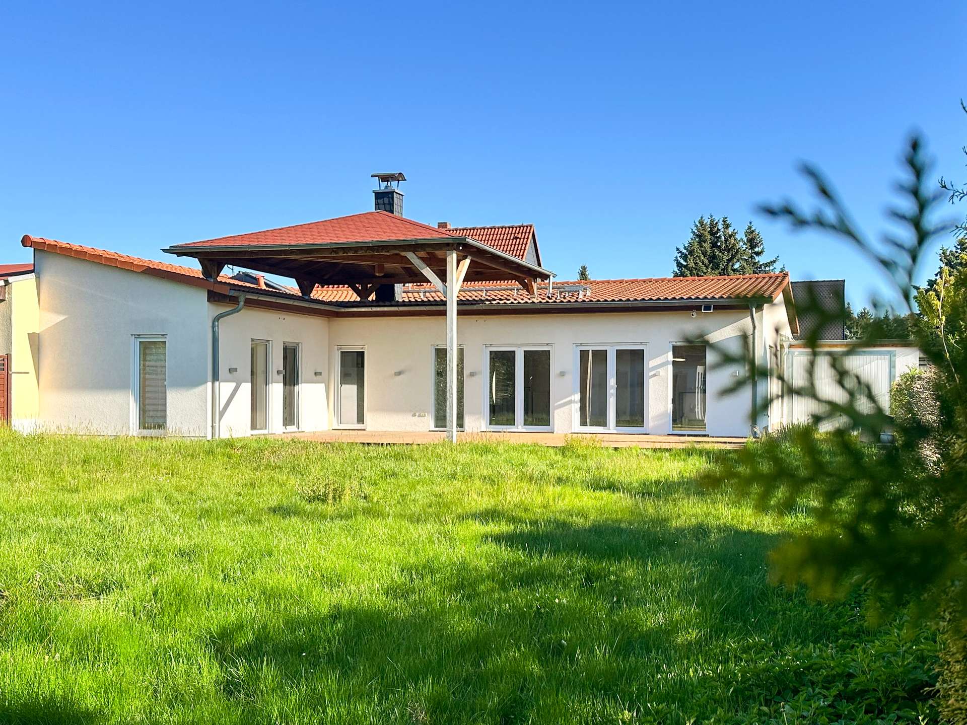 Single-story white house with a red-tiled roof and large glass doors, set in a sunny green yard under a clear blue sky.