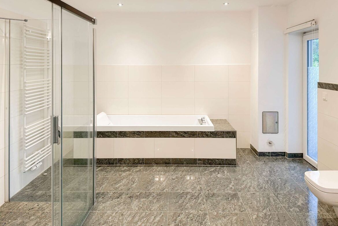 Modern bathroom with a built-in tub along a tiled wall, glass shower enclosure on the left, and a window on the right.